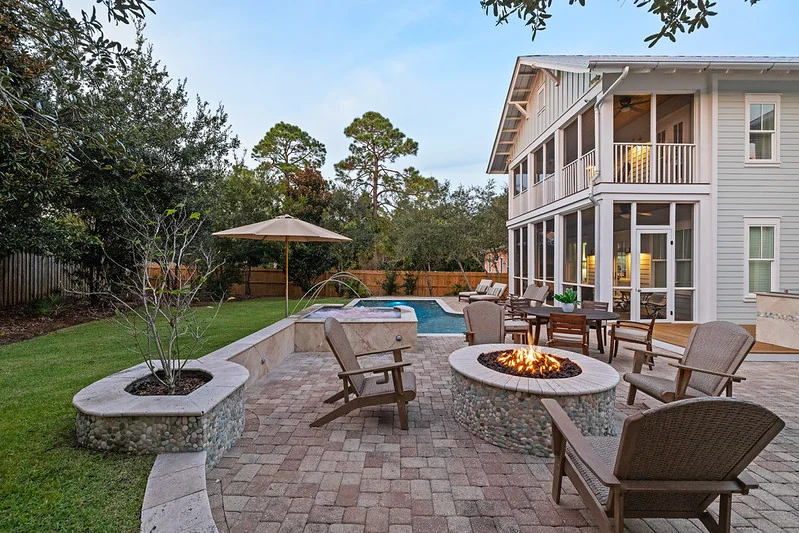 Outdoor entertaining at dusk: 12-seat covered lanai dining, pool-deck fire-bowl lounge, summer-kitchen grill counter.