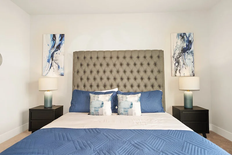 Queen guest bedroom: layered tailored coverlet, brass accent lamp, three-frame gallery above headboard, woven bench.